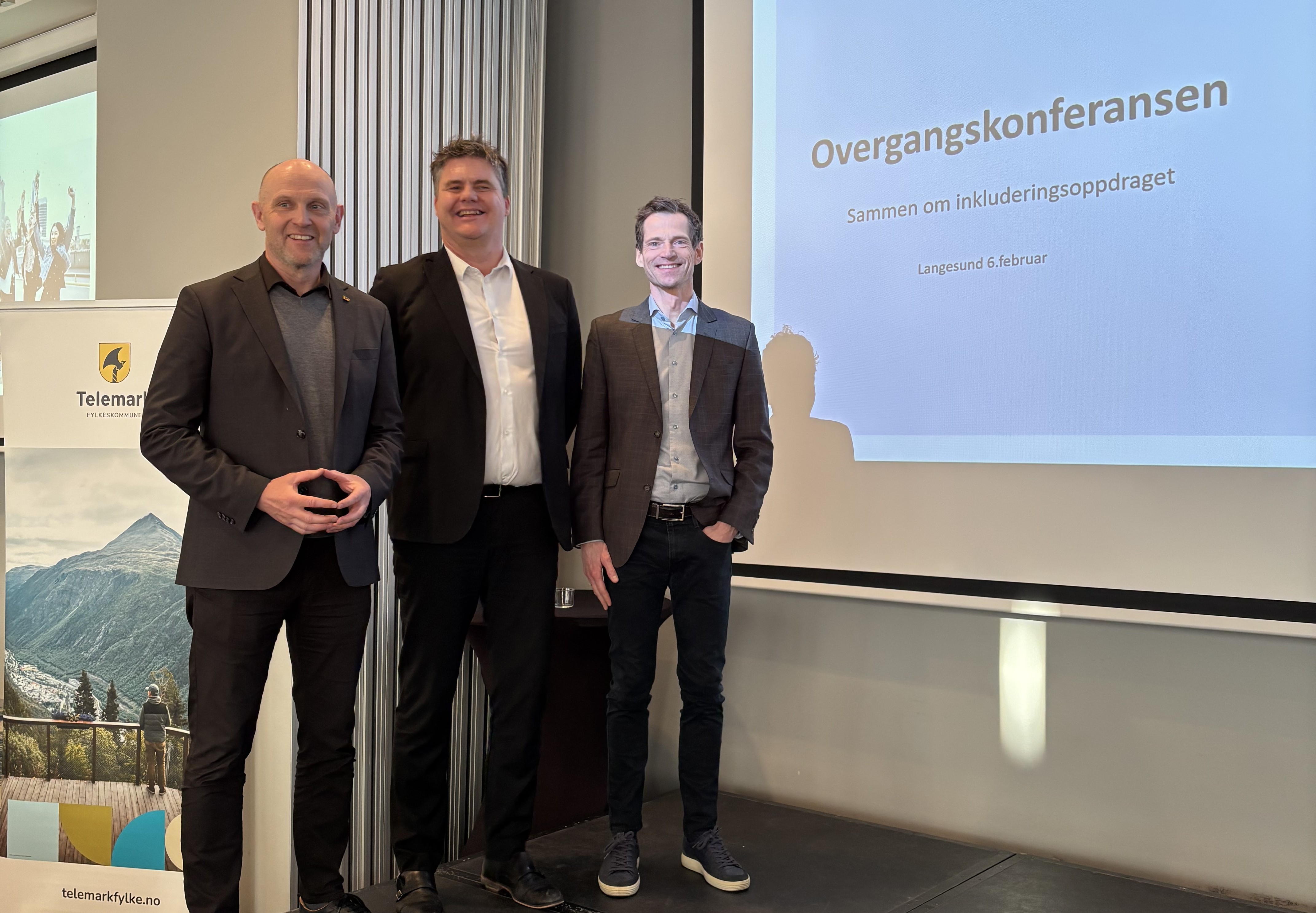 Tre menn i dress som står på en scene og smiler