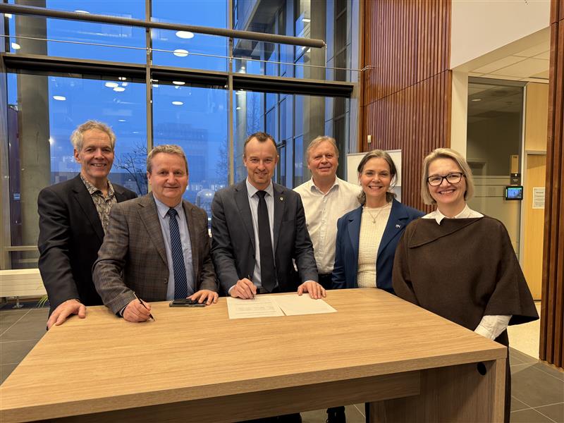 Bilde av seks personer, fire menn og to kvinner, som står ved et høyt bord og smiler til kamera.