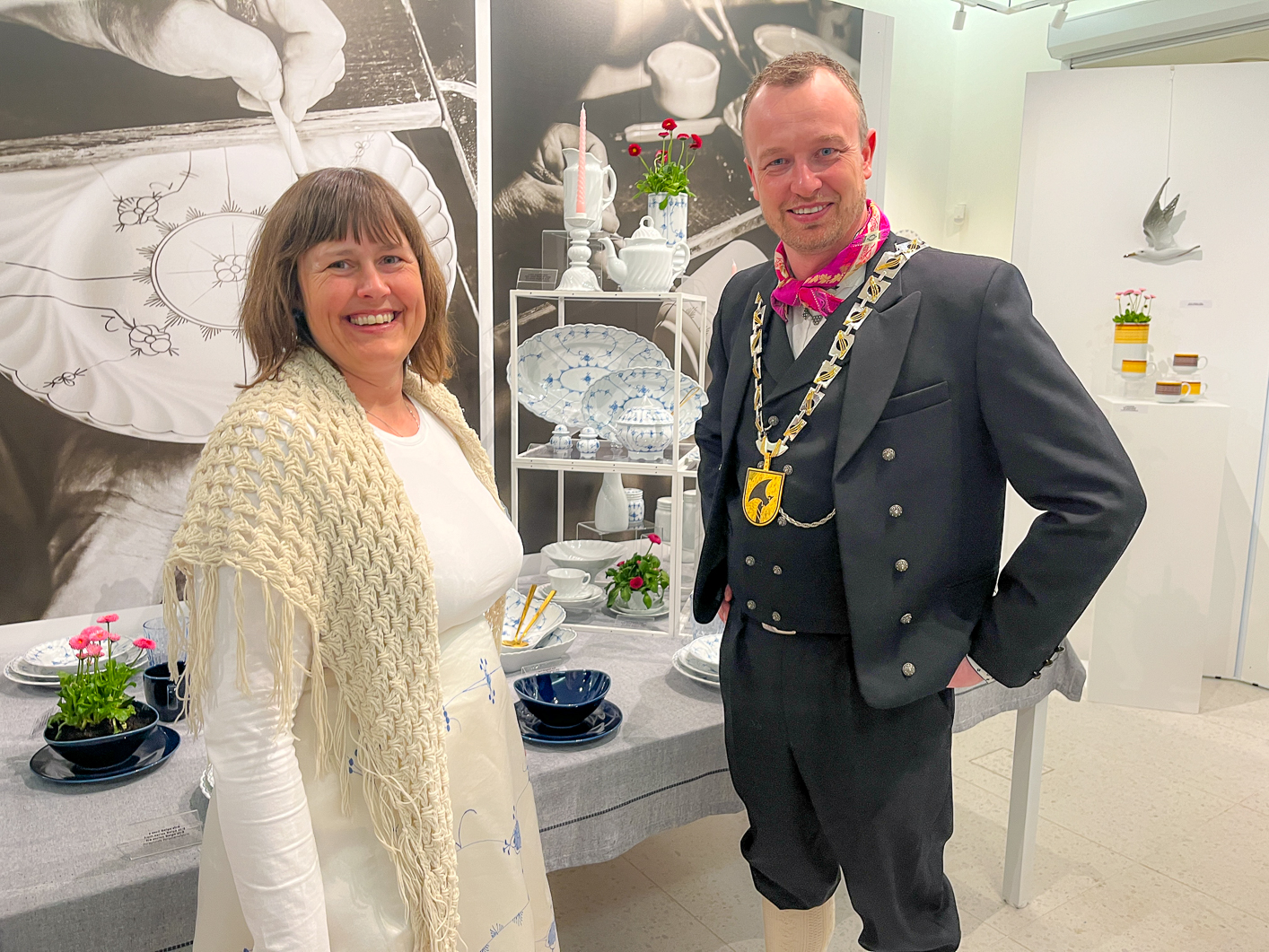 Mann og kvinne i bunad som står foran et bord med utstilling av ulike porselensgjenstander.