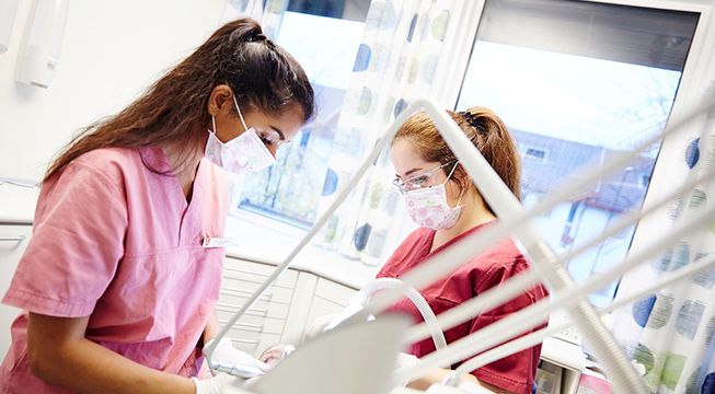 To kvinner med munnbind og helseuniform som jobber over en som ligger i en tannlegestol