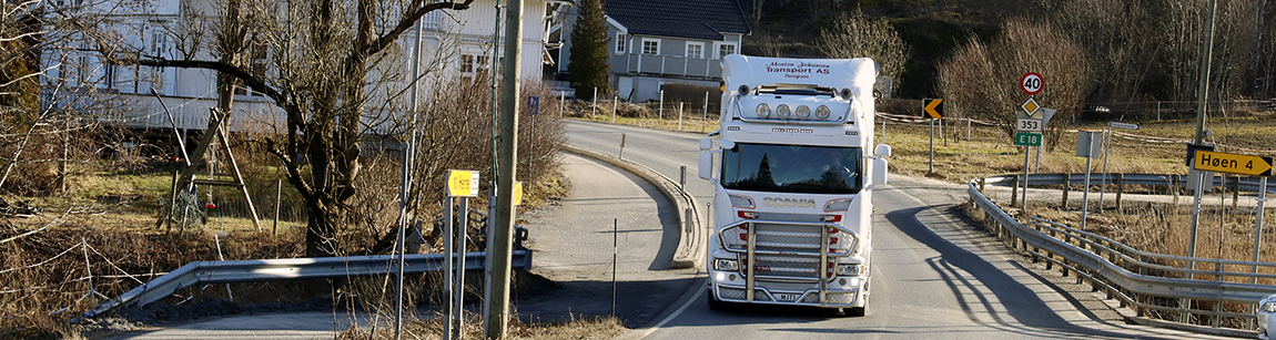 Viser smal vei på Rugtvedt, med uoversiktlig kryss inn fra høyre. Trailer kjører mot betrakteren av bilde. I bildets bakkant ser man bebyggelsen i området.