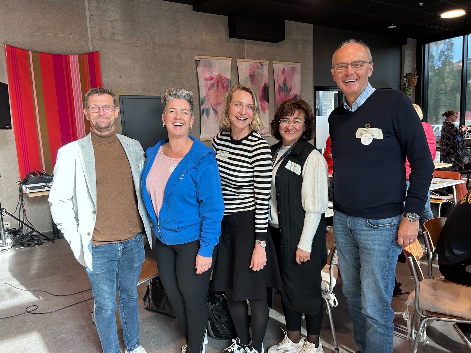 Fem mennesker, to menn og tre kvinner, står ved siden av hverandre og smiler mot kamera. Foto.