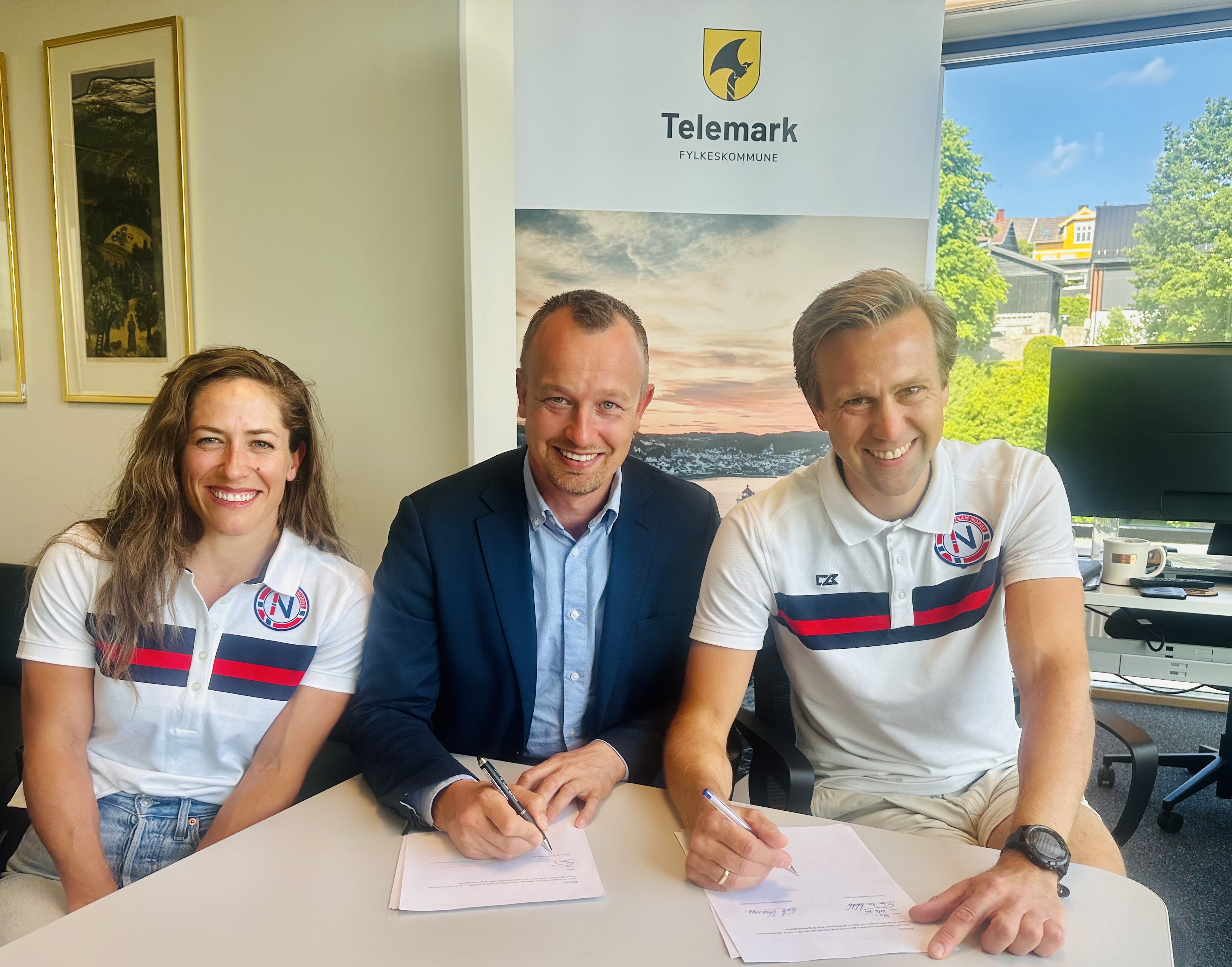 Ei kvinne og to menn smiler og skriv på eit papir. Dei signerer ein kontrakt.