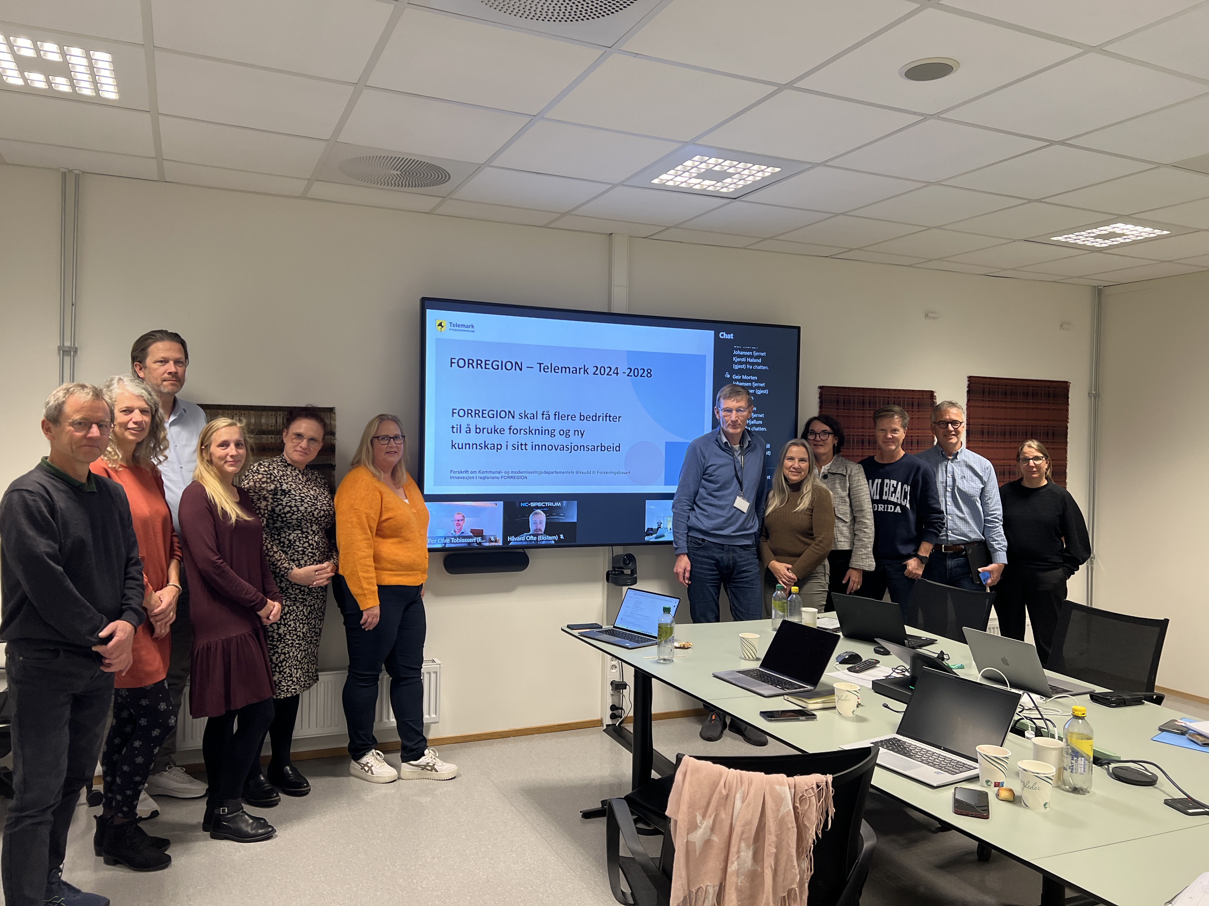 Bilde av flere mennesker i et rom som står samlet ved en skjerm.