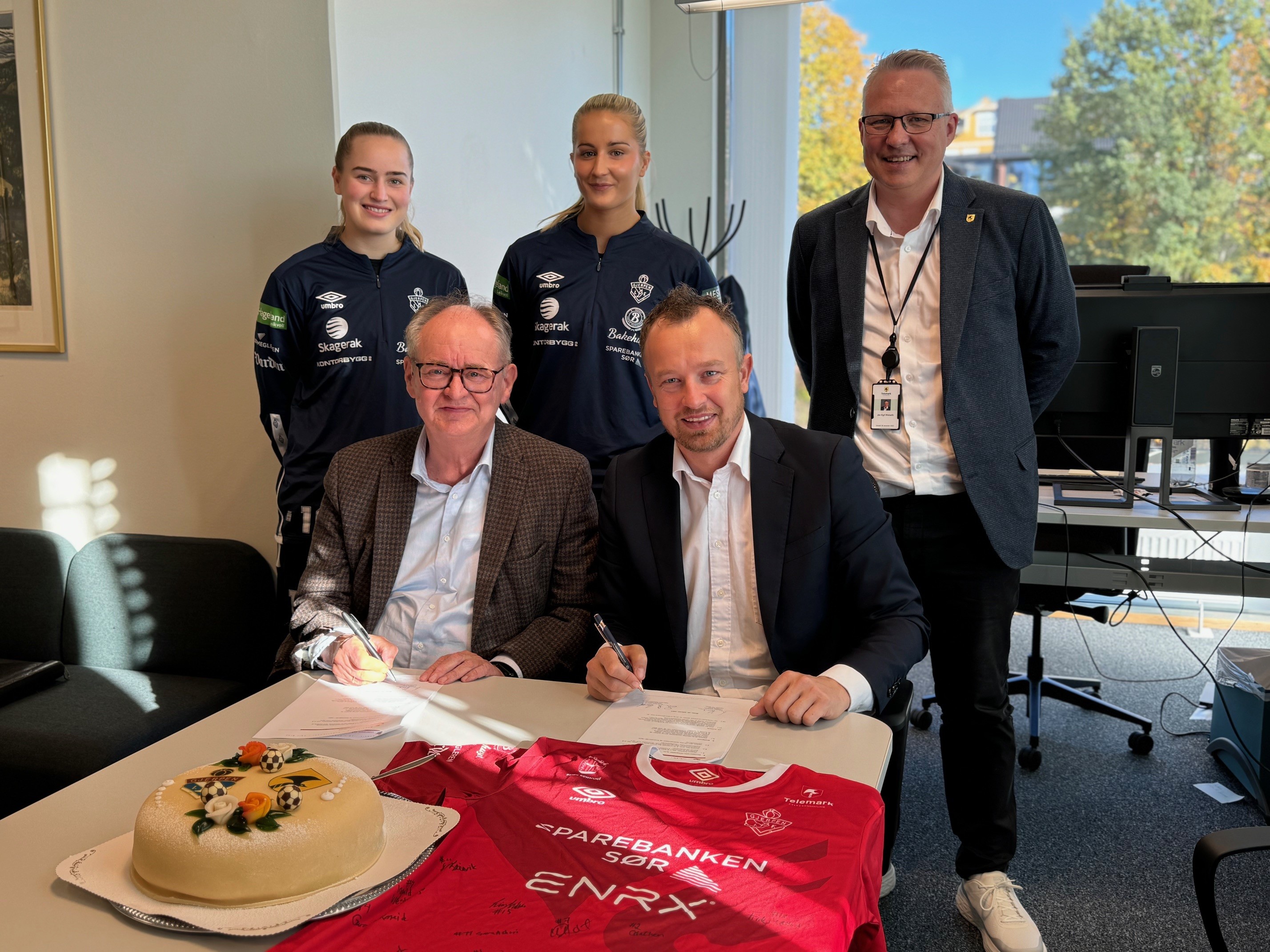 To kvinner i treningstøy og tre menn i dress står ved et bord med et ark på bordet som blir signert.