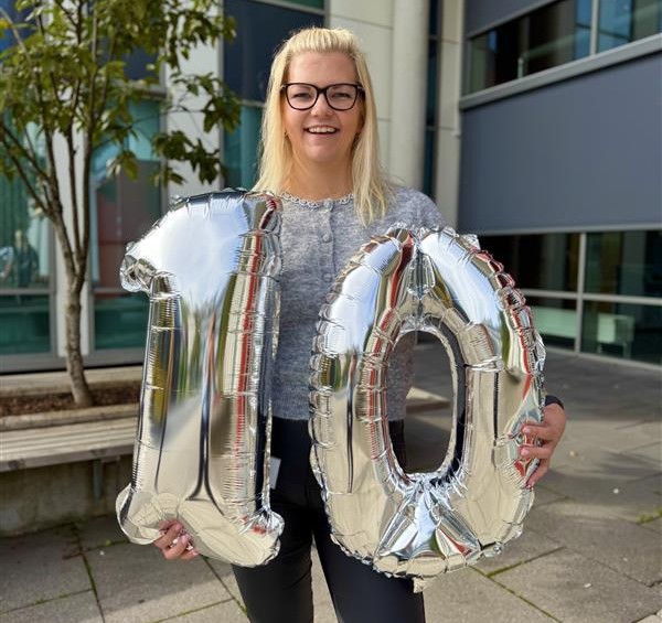 Kvinne som smiler til kamera og holder tallet ti opp i sølvfargede ballonger