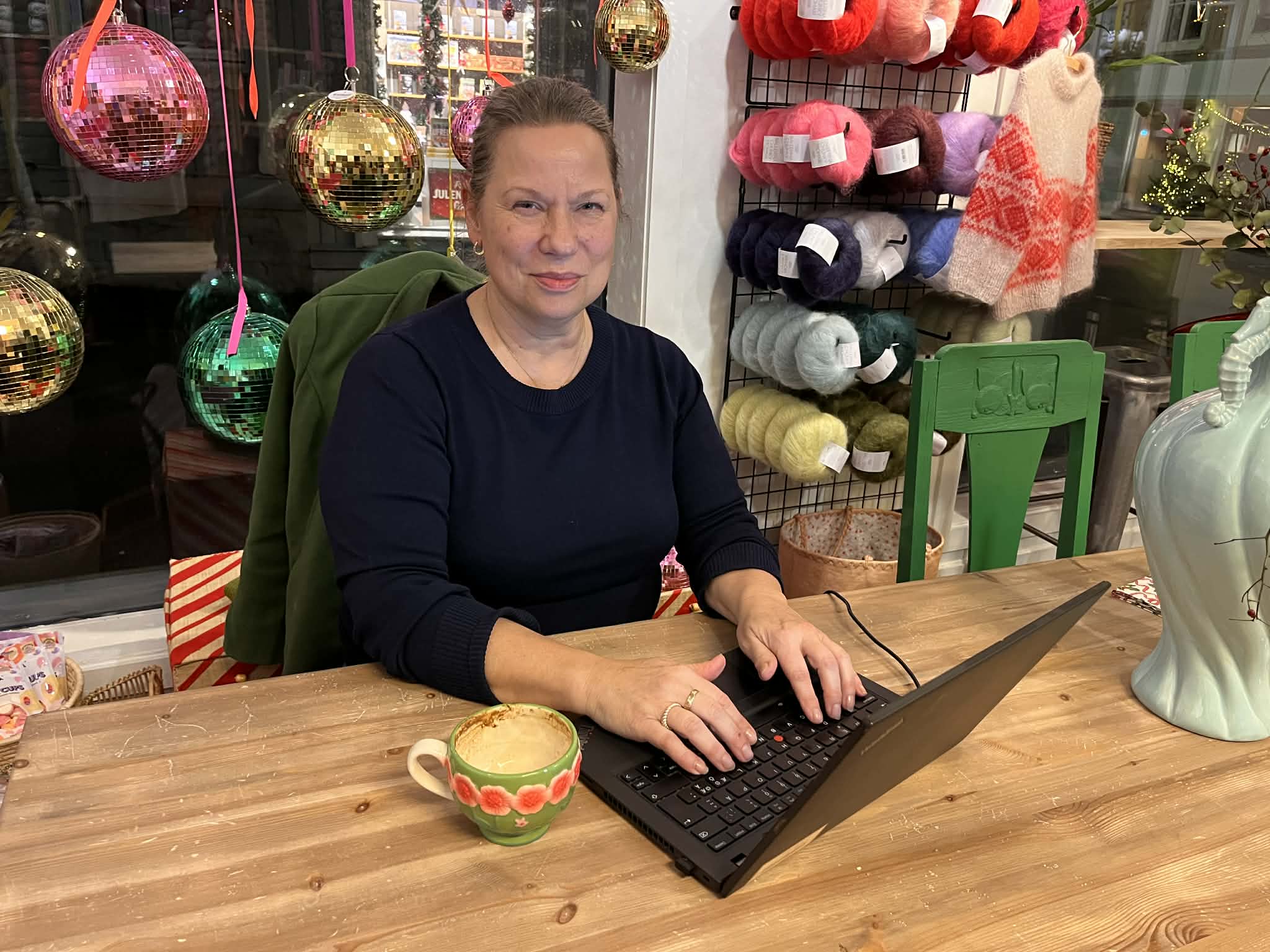 En kvinne som sitter bak et bord med en pc foran seg. Hendene hennes ligger på tastaturet og hun ser i kamera.