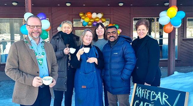Seks personer foran et bygg med ballonger på døra. Tre av personene holder en kaffekopp og alle smiler.