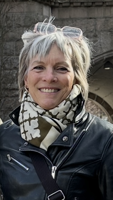 En smilende kvinne med kort, lyst hår og solbriller på hodet, iført en svart skinnjakke og et mønstret skjerf i grønt og hvitt. Hun står foran en steinbygning og ser rett inn i kameraet.