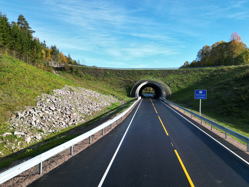 nnkjøringen til Skaravegtunnelen, en tofelts vei med gule og hvite vegmerker som leder inn mot en betongportal. På høyre side står et blått tunnelskilt med teksten «Skaravegtunnelen». Grønne skråninger og høstfargede trær omgir tunnelåpningen under en blå himmel.