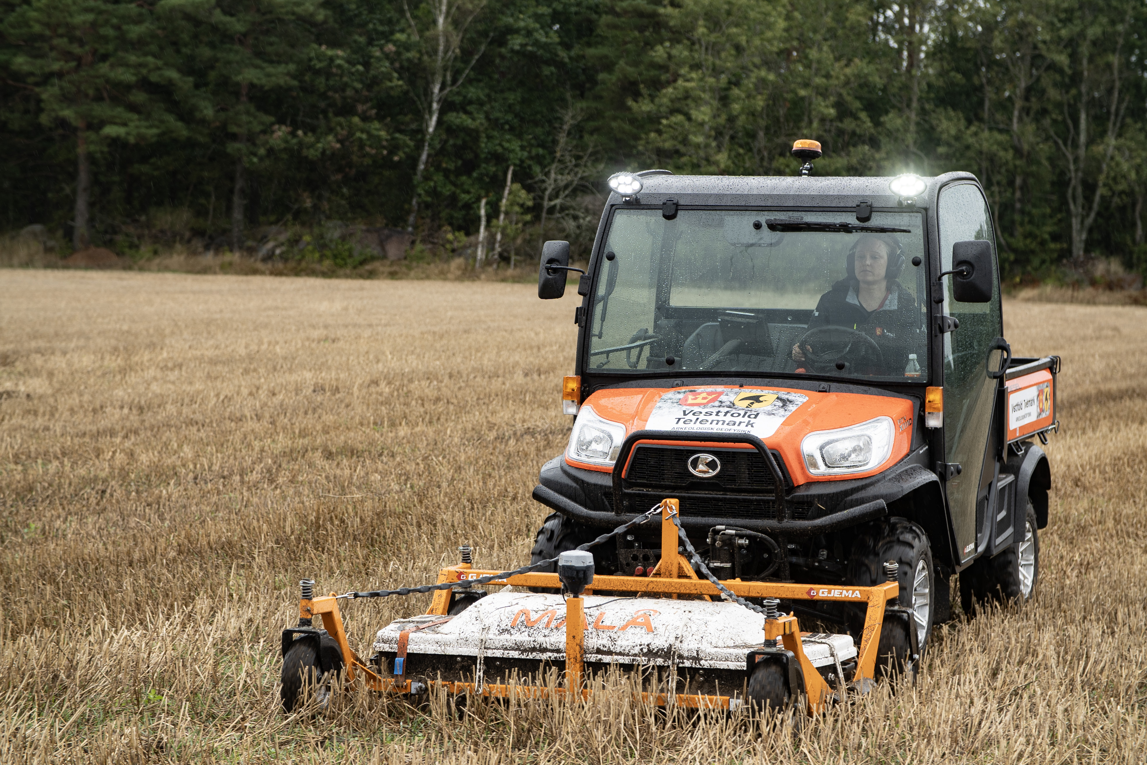 En person kjører en oransje Kubota nyttekjøretøy med Vestfold og Telemark fylkeskommune-logo over et tørt jordstykke, med en GJEMA Mila frontmontert slåmaskin.