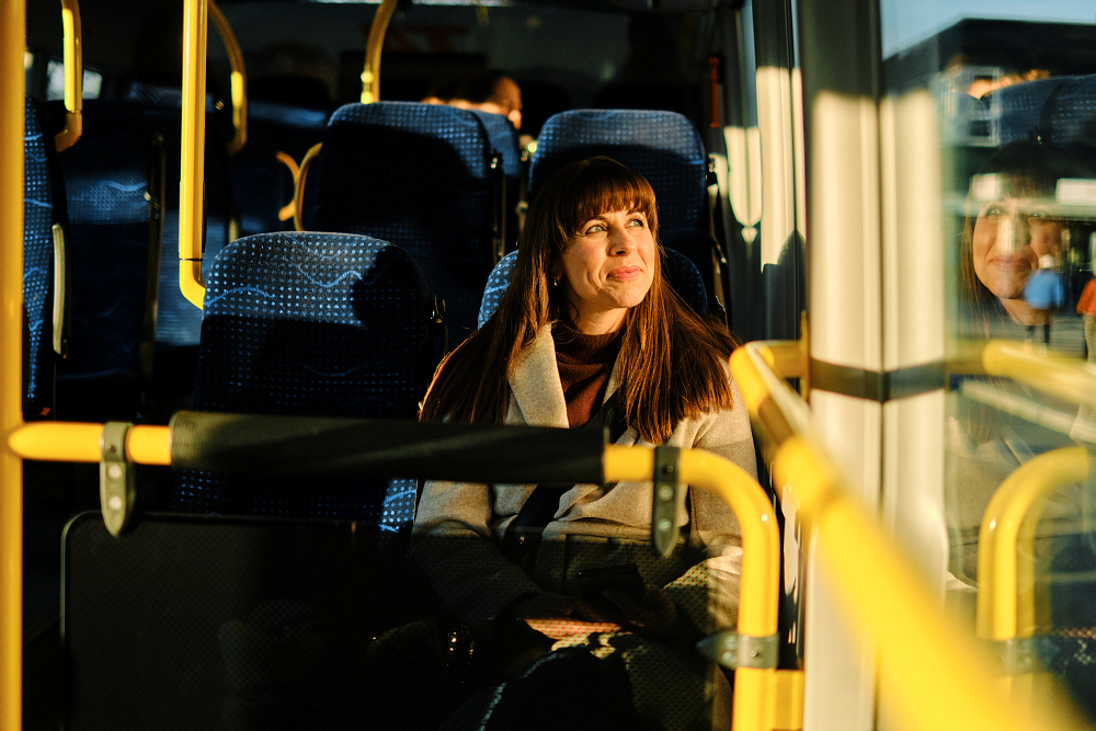 Et portrett av en kvinne som sitter på et offentlig transportmiddel, sannsynligvis en buss eller trikk. Hun har mørkt, halvlangt hår, en beige trenchcoat og et mørkt skjerf. Hun ser ut til å være i egne tanker, med blikket vendt litt nedover. Bak henne skimtes andre passasjerer og gule håndtak i vinduet. Bildet er tatt i dagslys, men med en litt dempet belysning inne i kjøretøyet.